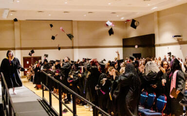 A large group of graduates in black caps and gowns joyfully toss their mortarboards into the air during a graduation ceremony. One graduate is walking across the stage while the audience and fellow students cheer and celebrate.